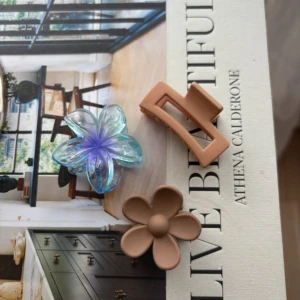 Hårklämmor i olika färger och former - Tre hårklämmor i olika stilar. En genomskinlig blomformad klämma med blå och lila nyanser, en beige klämma i form av en blomma, och en enkel beige klämma med rektangulär form. Perfekta för att sätta upp håret med stil.