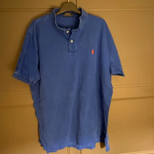Blå pikétröja från Ralph Lauren - Säljer en klassisk blå pikétröja från Ralph Lauren med den ikoniska loggan broderad i rött på bröstet. Tröjan har korta ärmar och knappar vid kragen. Perfekt för en stilren och avslappnad look.
