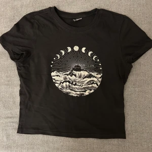 Svart t-shirt med tryck från H&M Divided - Säljer en svart t-shirt från H&M Divided med ett coolt tryck av månfaser och vågor på framsidan. T-shirten har korta ärmar och en rund halsringning. Perfekt för en avslappnad stil.