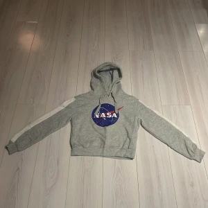 Grå NASA hoodie från Divided - Säljer en grå hoodie från Divided med NASA-logga i blått och rött på framsidan. Tröjan har en huva och vita ränder längs ärmarna. Bra skick i hoodien och det är en XS som passar 160! Perfekt för dig som gillar rymdtema och bekväma kläder! 🌌