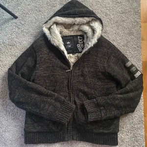 Fodrad cardigan - Säljer en mörkgrå jacka från med mjukt fuskpälsfoder och huva. Jackan har dragkedja framtill och ribbade muddar vid ärmslut och nederkant. Detaljer på ärmen med text och årtal. Perfekt för kyliga dagar! Skick 9/10. Skriv för fler frågor eller bilder. Priset är ej hugget i sten så kom med prisförslag.
