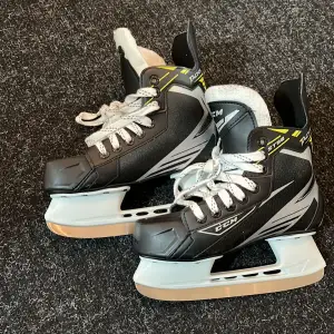 Säljer ett par svarta skridskor från CCM med vita snören och grå detaljer. De har en robust design med vadderad insida för extra komfort. Perfekta för isaktiviteter och har en stilren look.