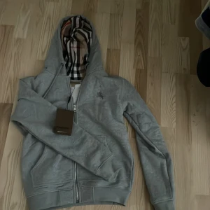 Burberry’s zip hoodie  - Säljer en snygg grå huvtröja från Burberry med klassiskt rutigt mönster i huvan. Tröjan har dragkedja och en diskret broderad logga på bröstet. Perfekt för en stilren look.