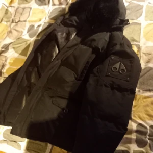 Svart dunjacka från Moose Knuckles - Säljer en svart dunjacka från Moose Knuckles med pälskrage och snygga detaljer på ärmen. Jackan har en quiltad insida och är perfekt för kalla dagar. Den har en klassisk design med flera fickor och en logotyp på insidan.