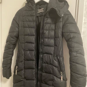 Svart dunjacka från Garoff - Snygg svart dunjacka från Garoff med huva och dragkedja framtill. Jackan har en quiltad design och praktiska fickor med dragkedjor. Perfekt för kyliga dagar när du vill hålla dig varm och stilren.