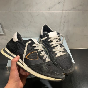 Svarta sneakers med vita detaljer - Snygga svarta sneakers från philippe model med vita snören och detaljer. Skorna har en klassisk design med en bekväm sula och en diskret orange detalj på sidan. Perfekta för en stilren look. Skick 8/10, bara lite smutsiga. Strl 43// skriv vid funderingar. Mvh