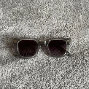 Grå solglasögon från Chimi 04 - Snygga grå solglasögon från Chimi med transparent båge och mörka linser. De har en fyrkantig form som ger en modern look. Perfekta för soliga dagar och kommer i ett stilrent vitt fodral.