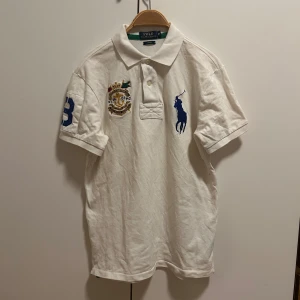 Vit pikétröja från Ralph Lauren - Säljer en klassisk vit pikétröja från Ralph Lauren med blå logotyp på bröstet och ett broderat emblem. Tröjan har korta ärmar och en krage med knappar. Perfekt för en stilren look!