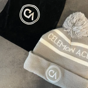 Grå mössa med tofs från Celemon Ace - Snygg grå mössa med vit rand och stor tofs på toppen från Celemon Ace. Mössan har en uppvikt kant med broderad logotyp framtill. Perfekt för kyliga dagar.