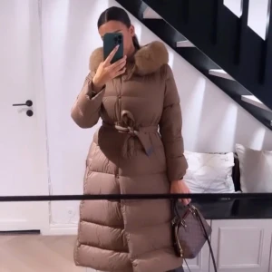 Brun dunjacka med pälsdetalj - Snygg brun dunjacka med pälsdetalj på kragen och knytbälte i midjan. Jackan har långa ärmar och en elegant, lång design som passar perfekt för kyliga dagar. Perfekt för att hålla värmen med stil!