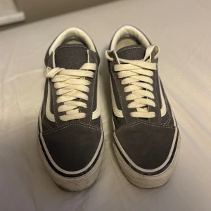 Grå och vita sneakers från Vans - Säljer ett par klassiska Vans sneakers i grått och vitt. Skorna har en ikonisk design med snörning och den typiska vågiga sidodetaljen. Perfekta för en avslappnad stil.