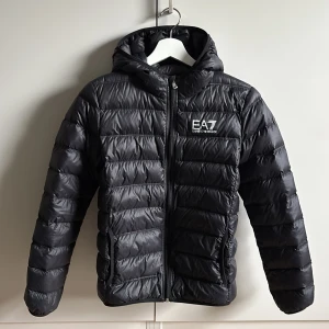 EA7 svart dunjacka  - Säljer en stilren svart dunjacka från Emporio Armani med huva och dragkedja. Använd men i jättefint skick, helt utan defekter.