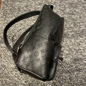 Svart ryggsäck med monogram - Snygg svart ryggsäck med monogrammönster. Den har en dragkedja och justerbara axelremmar. Perfekt för vardagsbruk eller resor. Stilren och praktisk design.