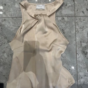 Beige ärmlös topp från Neo Noir - Snygg beige ärmlös topp från Neo Noir i ett glansigt material. Den har en elegant knutdetalj framtill som ger en stilren look. Perfekt för både vardag och fest!
