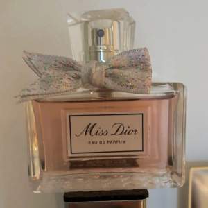 Säljer en elegant Miss Dior Eau de Parfum. Flaskan är rektangulär med en vacker, glittrig rosett på korken. Parfymen har en ljusrosa färg och ger en lyxig känsla. Perfekt för speciella tillfällen eller som en fin present.