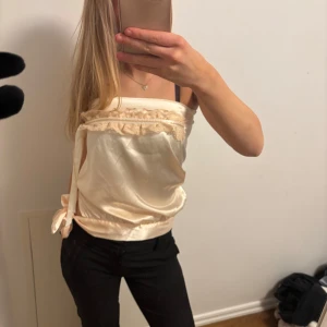 Beige satinlinne med spetsdetaljer - Säljer ett elegant beige linne i satin med vackra spetsdetaljer längs kanten. Linnet har en söt knytning på sidan och är perfekt för festliga tillfällen. Det är ärmlöst och har en mjuk, glansig yta som ger en lyxig känsla.