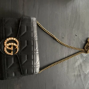 Svart quiltad axelväska med pärldetaljer - Snygg svart quiltad axelväska med guldkedja och pärlprydd logga. Väskan har en praktisk dragkedja inuti och flera fack för kort. Perfekt för både vardag och fest!