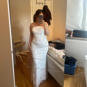 Vit axelbandslös långklänning - Säljer en elegant vit axelbandslös långklänning med vackra spetsdetaljer. Perfekt för fest eller bal! Klänningen har en figurnära passform som framhäver silhuetten. Den är i mycket bra skick.