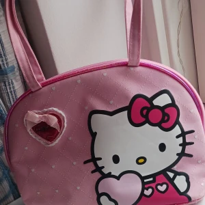 Rosa Hello Kitty handväska - Supergullig rosa Hello Kitty handväska med quiltat mönster och hjärtan. Väskan har en söt rosett och ett hjärta med Hello Kitty-logga. Perfekt för att ta med på en dag ute eller som en söt accessoar. Insidan är röd med små hjärtan. 🐱💖