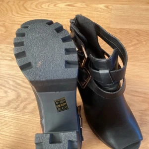 Svarta boots med klack och spännen - Snygga svarta boots i skinn med robust klack och coola spännen i metall. De har en dragkedja på sidan för enkel på- och avtagning. Perfekta för höst och vinter, dessa boots ger en edgy touch till vilken outfit som helst. Storlek 39.
