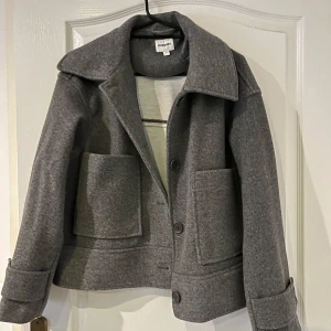 Grå kappa från Kapital - Säljer en snygg grå kappa från Kapital i mycket bra skick. Den har en klassisk design med stora fickor och knappar framtill. Perfekt för höst och vår, och den är superbekväm att ha på sig. Passar till både vardag och lite finare tillfällen.