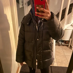 Svart pufferjacka med huva - Säljer en svart pufferjacka i mycket bra skick. Den är perfekt för vintern med sin varma vaddering och har en praktisk huva. Jackan har långa ärmar och stängs med både dragkedja och knappar. Perfekt för kalla dagar! Nypris 1300kr, köpte förra veckan och inte använd