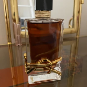 Ysl parfym libre intense eau de parfume  - Säljer en elegant parfymflaska från Yves Saint Laurent. Flaskan har en fyrkantig form med en mörk vätska inuti och en stilren svart kork. Den är dekorerad med en lyxig gulddetalj som ger en exklusiv känsla. Perfekt för den som vill ha en doft med stil och klass.  Parfymen är knappt använd bara 1-2 gånger! Det enda är att jag råkat tappa den så märket har ramlat av. Men innehållet är det samma! Den är 90ml brukar kosta annars 1500kr!