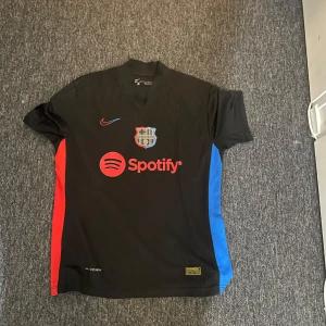 Svart FC Barcelona fotbollströja - Säljer en svart FC Barcelona tröja med korta ärmar. Den har röda och blå detaljer på sidorna och ett tryck med 'Lewandowski 9' på ryggen. Framtill finns Spotify-loggan och klubbmärket. Tröjan är i Dri-FIT material, perfekt för träning eller match!