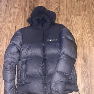 Svart dunjacka från Sail Racing - Säljer en svart dunjacka från Sail Racing i storlek S men passar även M.  Jackan är perfekt för kalla dagar med sin pufferstil och långa ärmar. Den har en praktisk dragkedja och en mysig luva man kan även vika in luvan så man kan använda jackan utan luva. Passar både till vardags och för utomhusaktiviteter. Håll dig varm och stilren i vinter! 🖤Nypris 4200kr kan även ta emot byten 