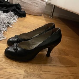 Svarta pumps med rosett - Säljer ett par eleganta svarta pumps  med en söt rosett på tån. De har en klassisk klack och är perfekta för både fest och mer formella tillfällen. Skorna är i bra skick och har en tidlös design som passar till det mesta.