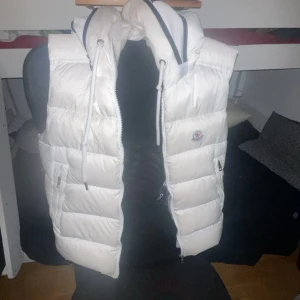 Vit dunväst med huva - Säljer en snygg vit dunväst från Moncler i mycket bra skick. Västen har en dragkedja framtill och en mysig huva med logotypdetaljer. Perfekt för kyliga dagar och ger en stilren look. Passar bra till både vardag och fest!