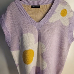 Lila stickad väst med blommor från SHEIN - Superfin lila stickad väst från SHEIN med stora vita blommor och gula detaljer. Den har en v-ringad design och är perfekt för vår och sommar. Västen är i bra skick och passar perfekt för en avslappnad och trendig look. 🌸