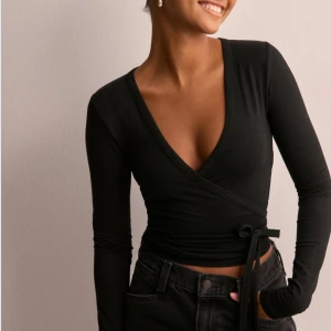 Svart omlottopp - Snygg svart omlottopp med lång ärm och djup v-ringning. Perfekt för både vardag och fest! Den har en tight passform som framhäver figuren och knytning i sidan för extra stilpoäng. Helt ny med prislapp! Passade tyvärr inte mig