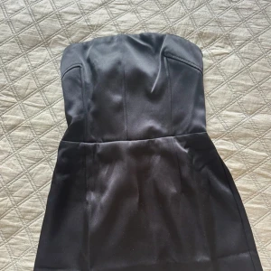 Svart axelbandslös klänning - Snygg svart axelbandslös klänning i satinmaterial. Perfekt för festliga tillfällen med en elegant och stilren design. Klänningen har en tight passform och en smockad rygg för extra komfort. Ny med prislapp 