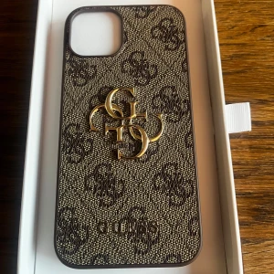 Mönstrat mobilskal från Guess - Säljer ett snyggt mobilskal (iPhone 14 och 13) från Guess med ett elegant mönster och guldiga detaljer. Skalet är i mycket bra skick (helt oanvänt pga köpt till fel iPhone modell) och har en stilren design som passar perfekt för att ge din mobil en lyxig touch. 