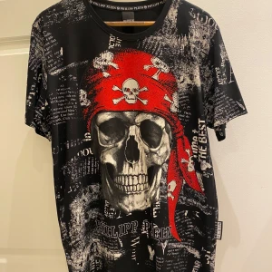 Philipp Plein T-shirt  - Svart t-shirt från Philipp Plein med ett stort dödskallemotiv och röd bandana. T-shirten har korta ärmar och är täckt av text och mönster i vitt och rött.