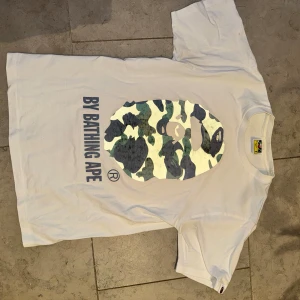 Vit t-shirt från A Bathing Ape - Säljer en vit t-shirt från A Bathing Ape med deras ikoniska kamouflagemönster i grönt och brunt på framsidan. T-shirten har korta ärmar och är i bra skick. Perfekt för en avslappnad stil eller streetwear-look. Storlek S enligt etiketten.
