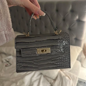 Grå krokodilpräglad handväska - Snygg grå handväska med krokodilpräglat mönster och gulddetaljer. Perfekt för att ge en lyxig touch till din outfit. 60kr