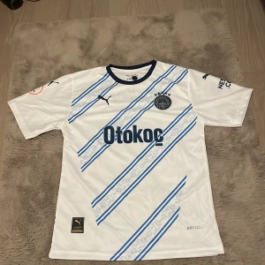 Vit och blå Fenerbahçe fotbollströja från Puma - Säljer en snygg vit fotbollströja från Puma med blå diagonala ränder och Fenerbahçe-logga. Tröjan har korta ärmar och är i mycket bra skick. Perfekt för fans eller samlare! Storlek 176, vilket motsvarar ungefär herr S.