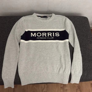 Grå tröja från Morris - Säljer en snygg grå tröja från Morris med texten 'Morris Rowing Club' i marinblått och vitt på bröstet. Tröjan är långärmad och har ribbade muddar vid ärmslut och nederkant. Perfekt för en stilren och avslappnad look!