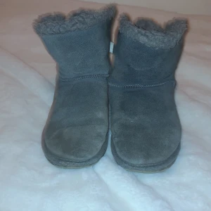 Grå uggs rosett - Säljer mysiga grå boots i mocka med fluffigt foder och söta rosetter bak. Perfekta för kalla dagar när du vill hålla fötterna varma och bekväma. De har en enkel design med en liten klack och är i bra skick. Passar perfekt till höst- och vinteroutfits! Använda i ett halvår, bra skick.