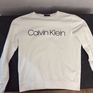 Vit tröja från Calvin Klein - Säljer en stilren vit tröja från Calvin Klein i storlek S. Tröjan har en klassisk design med långärmade ärmar och Calvin Klein-loggan tryckt på framsidan. Perfekt för en avslappnad och trendig look. Passar till både vardag och fest!