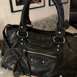 Svart skinnväska med dragkedja - Säljer en snygg svart skinnväska i mycket bra skick. Den har en cool design med dragkedja och silverdetaljer. Perfekt för både vardag och fest, i ganska bra skick. Bara att frpga om ni har funderingar den har små defekter på fragkedjornas band men annars i bra skick