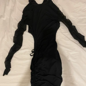 Svart långärmad klänning - Svart lång klänning med hål vid midjan som man kan dra ihopa för att de ska få de att se ut som att man har en mer tim glas figur den är även skrunahad vid rumpan så de ska se ut som att den är lite större har även dragkedja i ryggen 