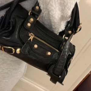 Svart axelväska med dragkedja - Snygg svart axelväska i skinn med guldfärgade detaljer och dragkedja framtill. Väskan har både handtag och en avtagbar axelrem, vilket gör den superpraktisk för alla tillfällen. Perfekt för både vardag och fest!