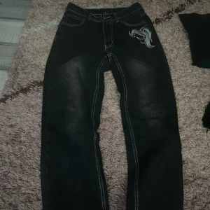 Svarta jeans med broderad detalj - Säljer ett par svarta jeans i bra skick med en cool broderad detalj på ena fickan. De har en loose passform och är perfekta för en avslappnad stil. Säljer för att den inte kommer till användning längre🫶