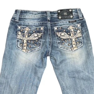 Miss Me Jeans - Snygga Miss Me Jeans med unika broderade detaljer på bakfickorna. De har en bootcut-stil som ger en retro vibe och är perfekta för både vardag och fest. Jeansen har en sliten look som ger dem en cool och avslappnad känsla. Perfekta för dig som vill sticka ut!
