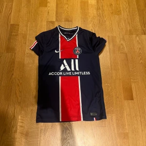 PSG fotbollströja från Nike - Säljer en snygg PSG fotbollströja i mycket bra skick. Den är mörkblå med en röd och vit rand i mitten. Tröjan är kortärmad och har Dri-FIT teknologi för bästa komfort. Perfekt för både träning och match! 