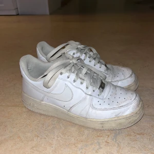 Vita Nike Air Force sneakers - Säljer ett par vita Nike Air Force sneakers. De är välanvända och har en del slitage, speciellt på insidan av hälen. Skorna har snörning och är i syntetmaterial. Perfekta för vardagsbruk eller träning.
