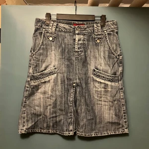 Jeanskjol med fickor och bälteshällor - Säljer en cool jeanskjol i en tvättad blå färg. Kjolen har två stora fickor fram och två mindre fickor bak, alla med snygga sömdetaljer. Den har också bälteshällor och en knappstängning fram. Perfekt för en avslappnad och trendig look!